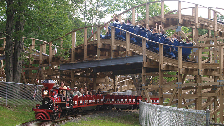 People in blue cars riding the Wooden Warrior roller coaster as people in red cars ride the Quassy Express Train